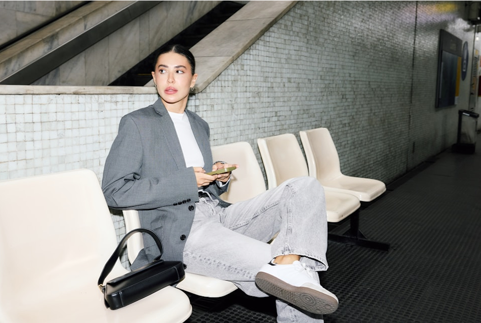 A person dressed in a gray blazer, white t-shirt, and light gray jeans relaxes on a row of white chairs in a tiled subway station, holding a phone with a black handbag placed beside them.