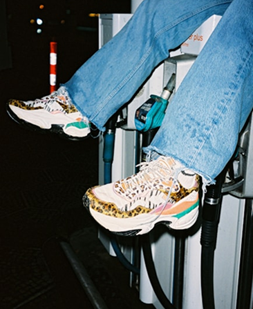 A person wearing light blue jeans and patterned sneakers sits atop a gas station pump at night, with fuel nozzles in the background and a street post visible nearby.